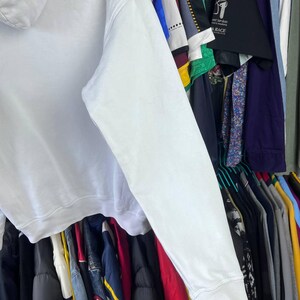 May include: A white hoodie with a long sleeve, seen from the side. The hoodie is made of a soft material and has a ribbed cuff. The background shows a rail of various clothing items.