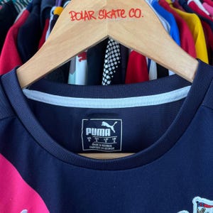 Pu&ograve; includere: Una maglia sportiva Puma blu navy con colletto bianco e dettagli rosa. La maglia ha il logo Puma e le informazioni sulla taglia stampate sul davanti. La maglia &egrave; appesa a un appendiabiti in legno con la scritta "POLAR SKATE CO.".
