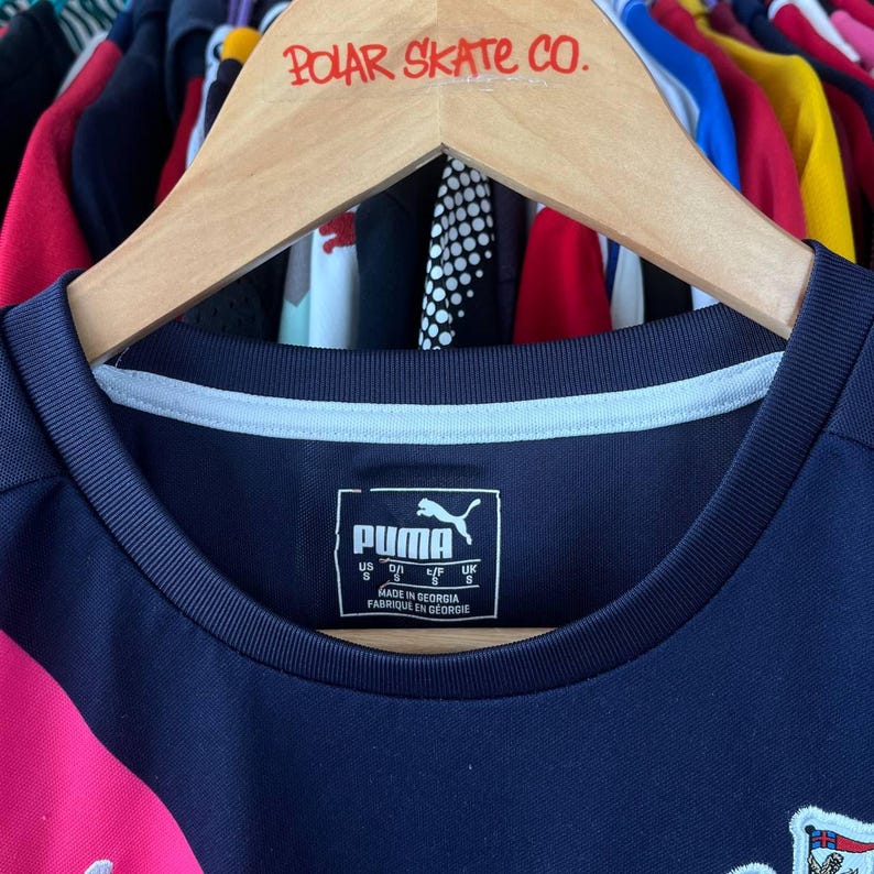 Pu&ograve; includere: Una maglia sportiva Puma blu navy con colletto bianco e dettagli rosa. La maglia ha il logo Puma e le informazioni sulla taglia stampate sul davanti. La maglia &egrave; appesa a un appendiabiti in legno con la scritta "POLAR SKATE CO.".
