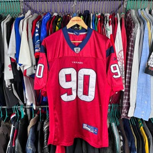 Pode incluir: Camisola de futebol vermelha com o número 90 na frente. A camisola tem o nome da equipa "Texans" na frente e o logotipo da Reebok na manga esquerda.