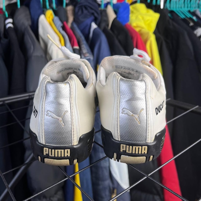 May include: Pair of white Puma sneakers with black soles. The heel features a silver panel with the Puma logo and the word "PUMA" in gold. The shoes are displayed against a backdrop of clothing.