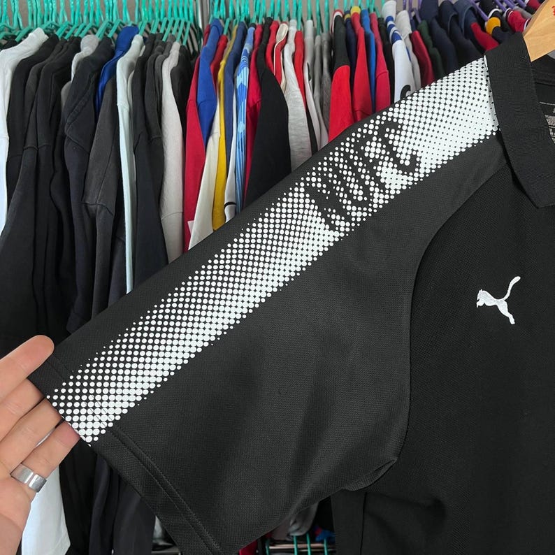 May include: Black Puma polo shirt with white dotted design on the sleeve and the word "UFC" printed on the sleeve. The Puma logo is on the chest. The shirt is hanging on a rack with other clothing.