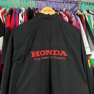 Puede incluir: Chaqueta negra con el logo rojo de Honda y el texto "The Power of Dreams". La chaqueta está colgada en una percha con otras prendas. La parte superior de la chaqueta tiene el texto "Polar Skate Co."