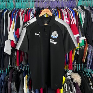 May include: Black short-sleeved polo shirt with white accents on the shoulders and sleeves. Features the Puma logo and a team crest on the chest. The shirt has a collared neckline and a button-down placket. The text "FUN88" is below the team crest.