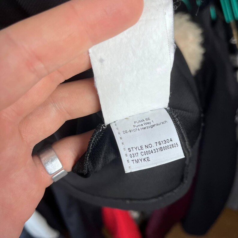 May include: Close-up of a white clothing tag with black text. The tag reads "PUMA SE Puma Way 1 DE-91074 Herzogenaurach STYLE NO. 751304 0317 C0004331B0002825 TMYKE". The tag is attached to a black garment.