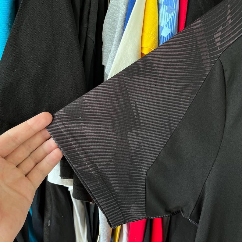 Puede incluir: Una camiseta negra de manga corta con un panel de patr&oacute;n gris oscuro distintivo en el hombro y la manga. La tela de la manga muestra una serie de l&iacute;neas finas, diagonales y curvas que crean un efecto visual sutil y texturizado sobre el cuerpo negro s&oacute;lido de la prenda.