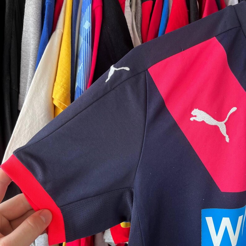 Pu&ograve; includere: Maglia sportiva blu navy con dettagli rossi. La maglia presenta un pannello rosa sulla spalla con logo Puma bianco. I polsini sono rossi e un logo blu e bianco &egrave; visibile.