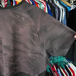 Puede incluir: Una camiseta de manga corta con un patr&oacute;n abstracto en gris oscuro y negro de l&iacute;neas finas, onduladas y paralelas. La prenda tiene un cuello redondo negro s&oacute;lido y un panel horizontal negro s&oacute;lido a lo largo del dobladillo inferior.