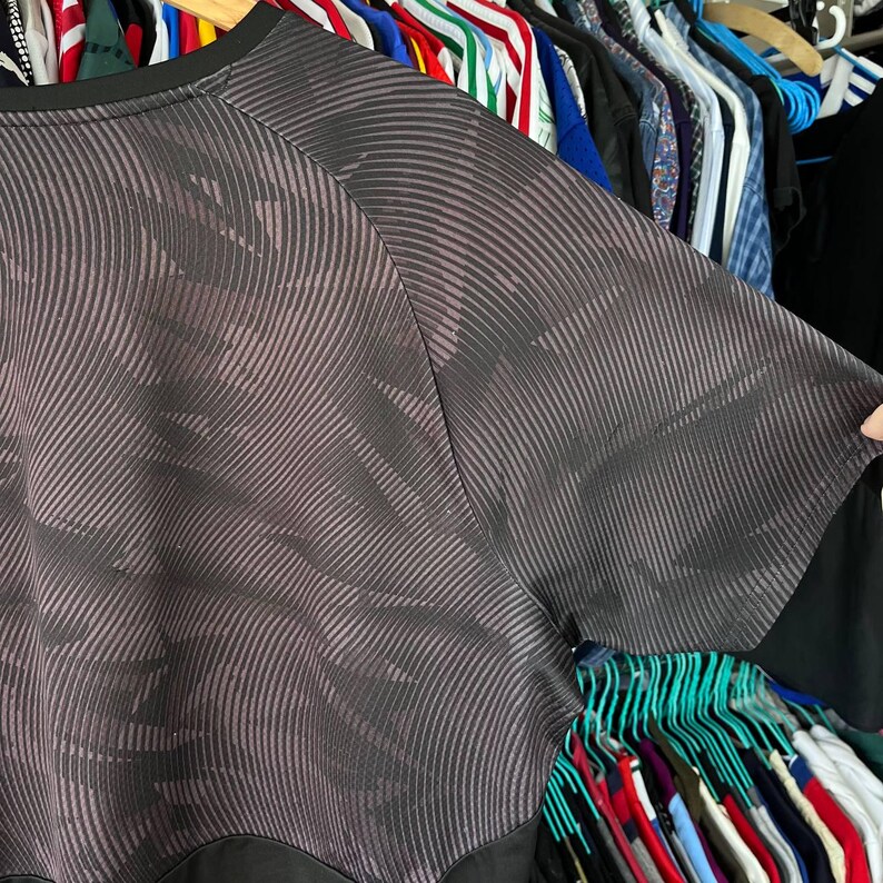 Puede incluir: Una camiseta de manga corta con un patr&oacute;n abstracto en gris oscuro y negro de l&iacute;neas finas, onduladas y paralelas. La prenda tiene un cuello redondo negro s&oacute;lido y un panel horizontal negro s&oacute;lido a lo largo del dobladillo inferior.