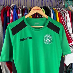 May include: Green Macron soccer jersey with black shoulder panels. The jersey features the Hibernian Football Club crest and the Macron logo. The jersey is hanging on a wooden hanger with the text "POLAR SKATE CO."