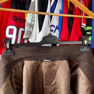 May include: A brown blazer with a small white grid pattern, hanging on a black hanger. The interior lining is a solid brown color. The blazer is part of a collection of clothing items on wooden hangers.