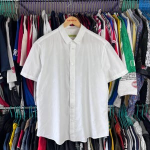 Puede incluir: Una camisa blanca de manga corta con botones, colgada de una percha de madera. La camisa tiene un cuello clásico y una botonadura frontal. El fondo muestra varias prendas de vestir en perchas.
