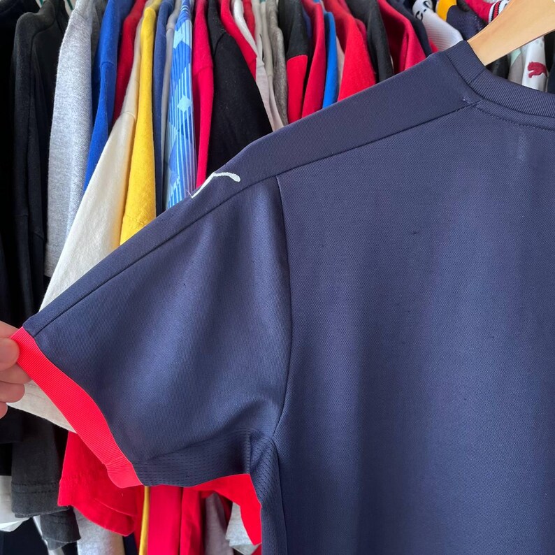 Pu&ograve; includere: Maglietta sportiva blu navy a maniche corte con finiture rosse sulle maniche. Un logo bianco &egrave; visibile sulla spalla. La maglietta &egrave; realizzata in un materiale leggero e traspirante.