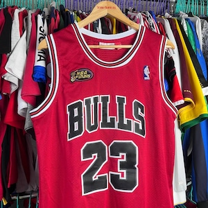 Puede incluir: Camiseta de baloncesto roja con "BULLS 23" en letras blancas y negras. Cuenta con un ribete blanco y negro alrededor del cuello y las sisas. Incluye un parche de "Finales de la NBA" y un logotipo de jugador de baloncesto.