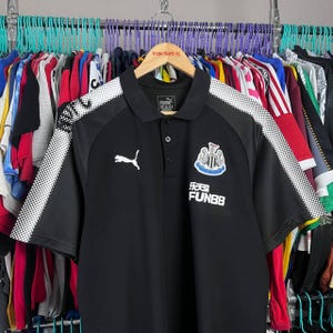 May include: Black Puma polo shirt with white accents on the shoulders and sleeves. Features the Newcastle United crest and FUN88 logo. The shirt is hanging on a wooden hanger with the text "YOUR SKATE CO."