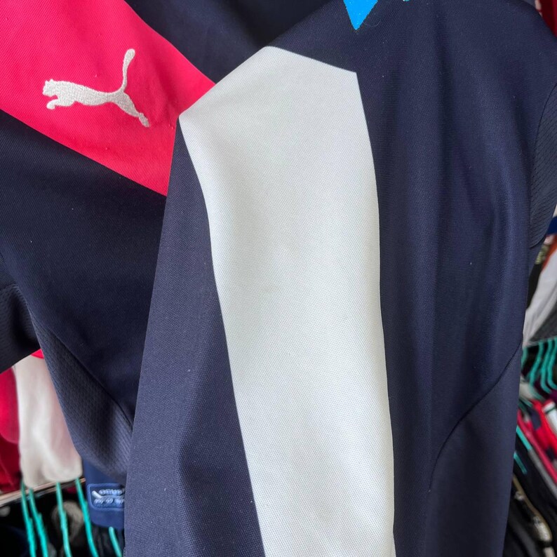 Pu&ograve; includere: Maglia sportiva blu navy con una striscia bianca sul lato e un pannello rosa sulla spalla. Un logo Puma argentato &egrave; visibile sul pannello rosa. La maglia ha una trama a rete sul lato.