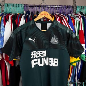K&ouml;nnte beinhalten: Ein dunkelgr&uuml;nes und schwarzes kurz&auml;rmeliges Sporttrikot mit V-Ausschnitt. Auf der Brust befindet sich ein wei&szlig;es Puma-Logo auf der rechten Seite, das Newcastle United-Wappen auf der linken Seite und der Text FUN88 mit chinesischen Schriftzeichen.