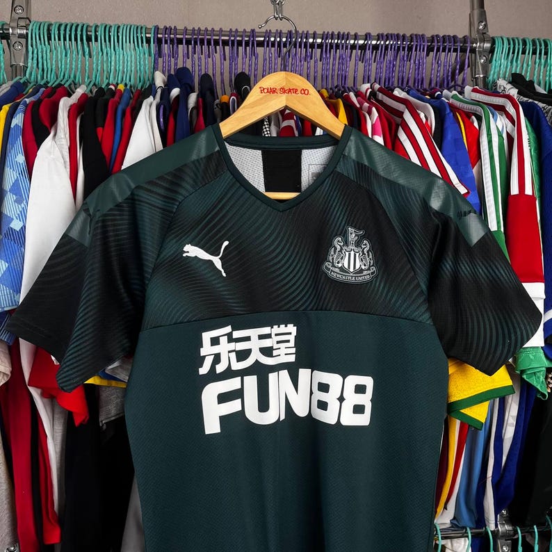 K&ouml;nnte beinhalten: Ein dunkelgr&uuml;nes und schwarzes kurz&auml;rmeliges Sporttrikot mit V-Ausschnitt. Auf der Brust befindet sich ein wei&szlig;es Puma-Logo auf der rechten Seite, das Newcastle United-Wappen auf der linken Seite und der Text FUN88 mit chinesischen Schriftzeichen.