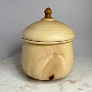 Puede incluir: Una caja de madera con tapa y un pomo en la parte superior. La caja está hecha de madera clara y tiene un acabado natural.