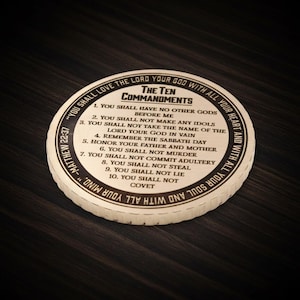 May include: A round, off-white coaster with the text "The Ten Commandments" in black. The text includes the ten commandments and a quote from Matthew 22:37. The coaster has a dark wood grain background.