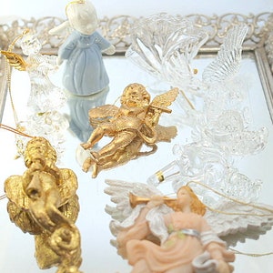 Pode incluir: Uma coleção de sete enfeites de anjo, incluindo um anjo de porcelana azul, um anjo de ouro e cinco anjos de vidro transparente. Os enfeites são exibidos em uma bandeja prateada com uma borda decorativa.