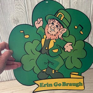 May include: A green shamrock-shaped cutout with a cartoon leprechaun wearing a green hat and holding a pot of gold. The leprechaun is dancing and there are musical notes around him. The cutout has the text "Erin Go Braugh" on a yellow banner at the bottom.