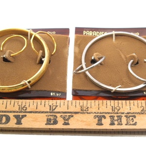 May include: Two pairs of hoop earrings, one gold and one silver, displayed on brown cards with the text "PARADISE DUO" and a price tag of $5.97. The earrings are shown on a wooden ruler.