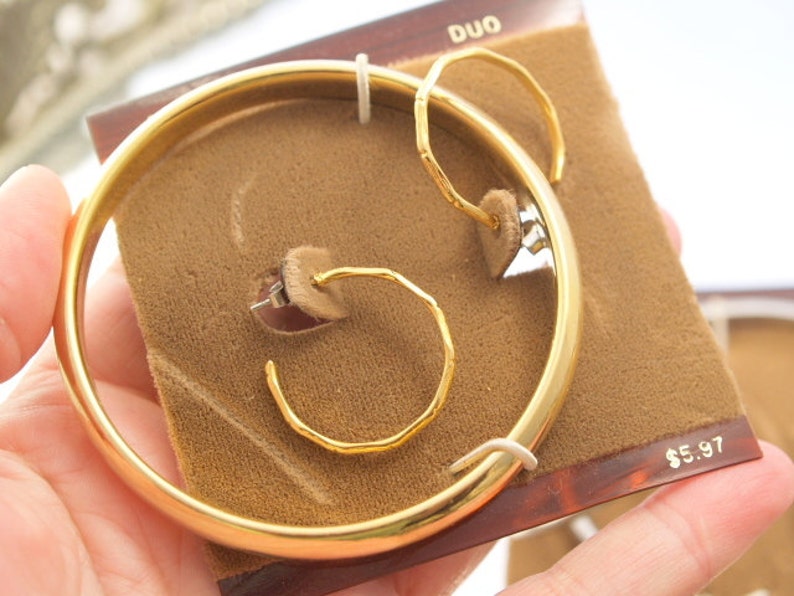 May include: Gold-toned jewelry set with a bangle bracelet and two pairs of hoop earrings. The set is displayed on a brown velvet background with the word "DUO" and the price "$5.97" printed on the packaging.