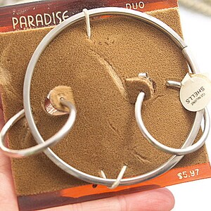 May include: A pair of silver hoop earrings on a brown velvet background. The earrings are labeled "Genuine Shells". The price is $5.97.