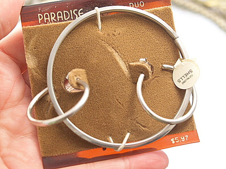 May include: A pair of silver hoop earrings on a brown velvet background. The earrings are labeled "Genuine Shells". The price is $5.97.