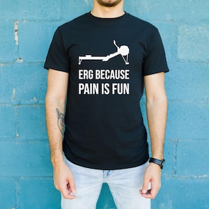 May include: Black crew neck t-shirt with the white text "ERG BECAUSE PAIN IS FUN" and a graphic of a rowing machine. The shirt is worn by a person in light blue jeans against a blue wall.
