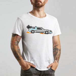 Puede incluir: Camiseta blanca con una imagen pixelada de un coche plateado con detalles azules y negros, llamas y una raya azul. El diseño del coche recuerda a un vehículo de película clásico. La camiseta es de cuello redondo.