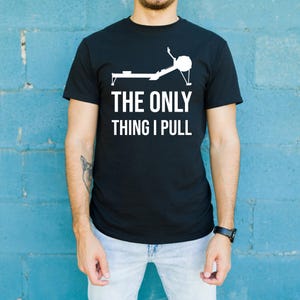 May include: Black crew neck t-shirt with the text "THE ONLY THING I PULL" in white. The shirt features a white graphic of a rowing machine. The person is wearing light blue jeans and a black watch.