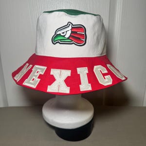 May include: A bucket hat featuring a white crown with a green top panel and a red brim. The front displays an embroidered eagle head logo in green, white, and red. The word MEXICO is stitched in large white block letters around the red brim.