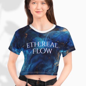 May include: A crop top with a blue and gold abstract design, featuring the words "ETHEREAL FLOW" in white. The shirt has a white collar and short sleeves. The fabric has a swirling, watercolor-like pattern.
