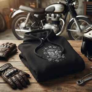 Pode incluir: Moletom preto com um gráfico de diagrama de motor branco, dobrado sobre uma superfície de madeira. Luvas de motociclista de couro marrom e um capacete de motocicleta vintage também são visíveis. Uma motocicleta clássica está ao fundo.