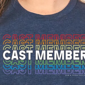 May include: A navy blue t-shirt with the text "CAST MEMBER" printed in white with rainbow colored outlines. The text is repeated five times, getting progressively smaller.