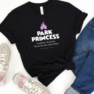 Może przedstawiać: Czarny T-shirt z grafiką r&oacute;żowego zamku i tekstem "Park Princess. Lead me. Feed me. Never let me wait in line."