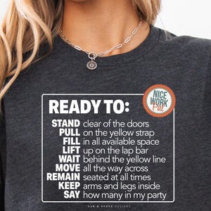 May include: A dark heather grey t-shirt with a white graphic that says "READY TO:" and lists a set of instructions. The instructions include "STAND clear of the doors", "PULL on the yellow strap", "FILL in all available space", "LIFT up on the lap bar", "WAIT behind the yellow line", "MOVE all the way across", "REMAIN seated at all times", "KEEP arms and legs inside", and "SAY how many in my party". The t-shirt also has a small white circle with the text "NICE WORK Pat" in blue and green.