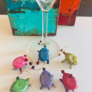 May include: Six colorful turtle-shaped wine glass charms. The charms are made of glass and have a gold-colored metal ring. The charms are arranged on a white surface.