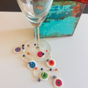 May include: Set of six colorful glass charms with a white background and a blue evil eye design. The charms are attached to gold rings and are perfect for marking wine glasses.