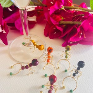 May include: Six colorful ant-shaped wine glass charms. Each charm is made of beads and wire and features a different color ant. The charms are on a white surface.