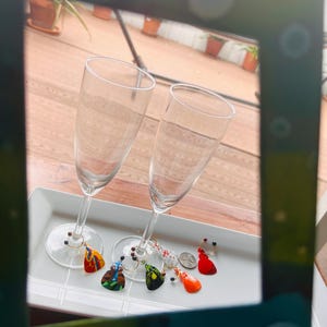 May include: Two champagne flutes with colorful glass charms hanging from the stems. The charms are shaped like guitar picks and feature different designs and colors.