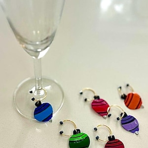 May include: Colorful wine glass charms in blue, red, orange, purple, and green. Each charm is shaped like a Christmas ornament with a gold-colored ring to attach to the glass. The charms are on a white surface next to a wine glass.