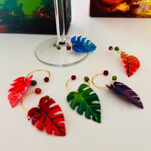 May include: A collection of colorful leaf-shaped wine charms. Each charm features a gold ring with a painted leaf in shades of red, green, blue, and purple. Small, round beads in complementary colors accent each charm, adding a decorative touch.
