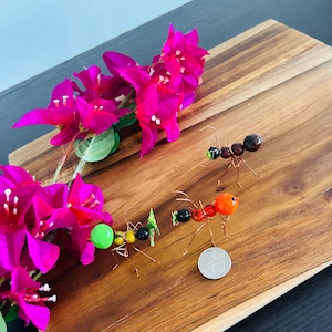 May include: Three colorful beaded ants made of glass and wire on a wooden surface with pink flowers. The ants are different sizes and have different colored beads. A US quarter is included for scale.