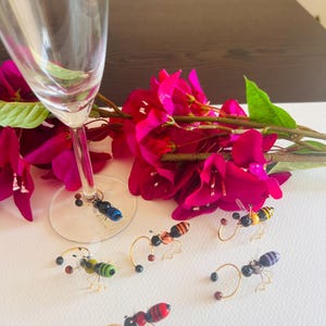May include: Six colorful glass insect wine glass charms. The charms are shaped like bees and ants and have gold wire legs. The charms are on a white surface with a glass of champagne and pink flowers in the background.