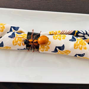 May include: A white napkin with a yellow and blue floral pattern is folded and held together with a decorative ant napkin ring. The ring is made of dark brown wood and has a small orange bead.