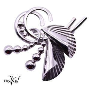 May include: A silver brooch with a geometric design featuring a series of curved lines, a chain of beads, and two fan-shaped elements.