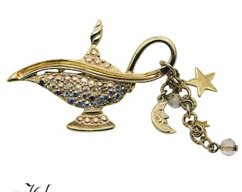 May include: Gold-toned Aladdin's lamp brooch with iridescent rhinestones and dangling charms. The charms include a gold star, crescent moon, and a clear bead. The brooch has a curved handle and a decorative base.